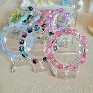 Op de afbeelding: Een verzameling kleurrijke armbanden met kralen, tentoongesteld op transparante standaards. De armbanden hebben verschillende kleuren, waaronder blauw, roze, paars en groen, met decoratieve bedels. De kralen variëren in grootte en textuur.