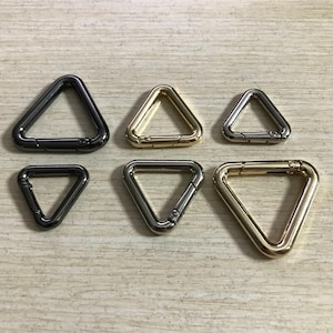 May include: Six metal triangle carabiner clips in various finishes: black, gold, and silver. The clips are arranged on a light wood-grain surface. These clips are designed for attaching straps or other accessories.