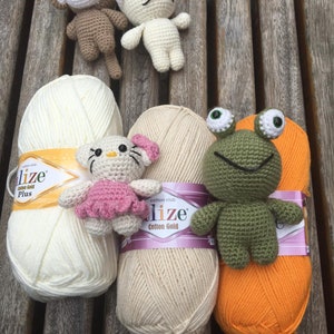 May include: A collection of handmade crochet toys, including a monkey, cat, frog, and a Hello Kitty-inspired character, displayed with skeins of cream, beige, and orange yarn. The toys are in various colors, and the yarn has gold and purple labels.