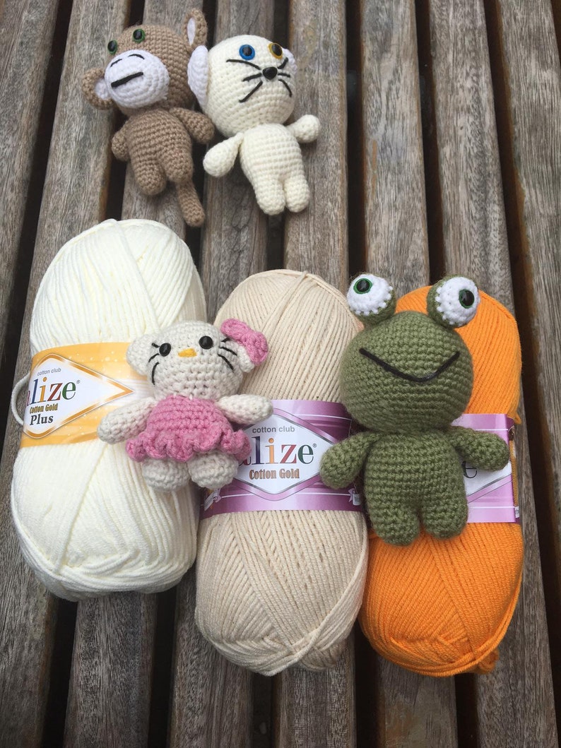 May include: A collection of handmade crochet toys, including a monkey, cat, frog, and a Hello Kitty-inspired character, displayed with skeins of cream, beige, and orange yarn. The toys are in various colors, and the yarn has gold and purple labels.