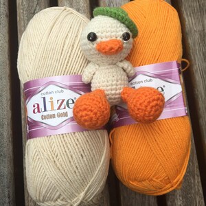 May include: A handmade crochet duckling with an orange beak and feet, wearing a green beret, sits between two skeins of yarn. One skein is cream-colored, and the other is bright orange. The yarn is labeled "Cotton Gold".