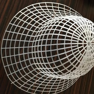 May include: White plastic mesh structure, possibly a plant support or decorative element. The mesh is constructed of a grid pattern, forming a curved, open shape. The object is set against a dark brown wooden surface.