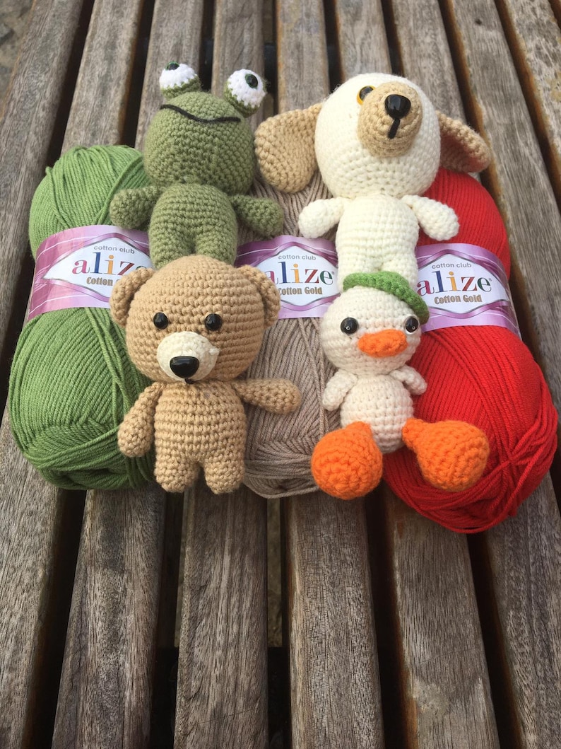 May include: A collection of handmade crochet toys, including a green frog, a beige bear, a white dog, and a white duck with orange feet. The toys are arranged with green, beige, and red yarn skeins.