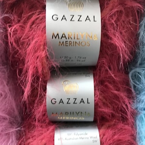 Puede incluir: Dos ovillos de hilo peludo rosa y azul etiquetados como "GAZZAL MARILYN& MERINOS" con el peso y el metraje indicados en la etiqueta.