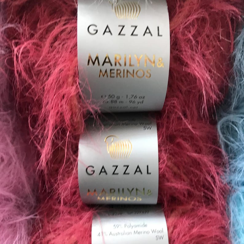 Puede incluir: Dos ovillos de hilo peludo rosa y azul etiquetados como "GAZZAL MARILYN& MERINOS" con el peso y el metraje indicados en la etiqueta.