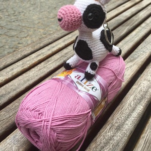 May include: A handmade crochet cow keychain with black spots, a pink nose, and small yellow horns, sitting on a ball of pink yarn. The yarn is labeled "Alize Cotton Gold". The cow is white with black spots and a silver keychain ring.
