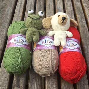 May include: A collection of handmade crochet toys, including a green frog and a white dog, displayed with three skeins of Alize Cotton Gold yarn in green, tan, and red. The yarn labels are visible.