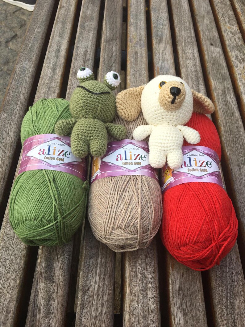 May include: A collection of handmade crochet toys, including a green frog and a white dog, displayed with three skeins of Alize Cotton Gold yarn in green, tan, and red. The yarn labels are visible.