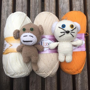 May include: A collection of yarn balls in cream and orange, with two crocheted animal figures. One is a brown monkey with a white face, and the other is a white cat with blue and yellow eyes. The yarn is labeled "Cotton Club".