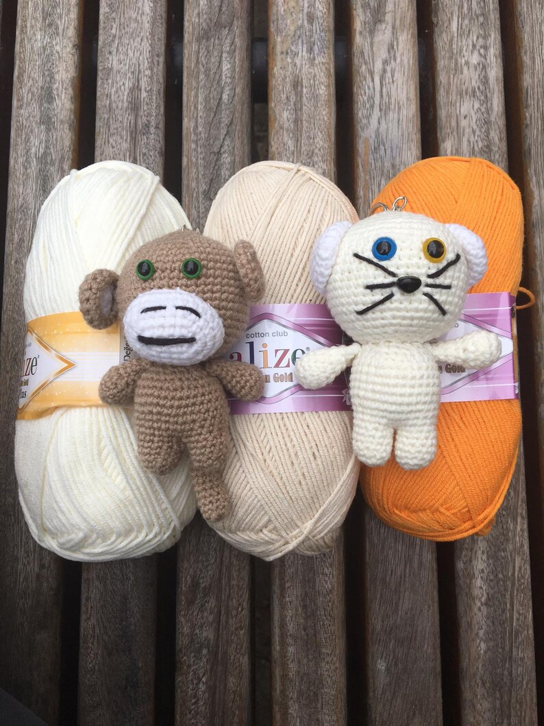 May include: A collection of yarn balls in cream and orange, with two crocheted animal figures. One is a brown monkey with a white face, and the other is a white cat with blue and yellow eyes. The yarn is labeled "Cotton Club".