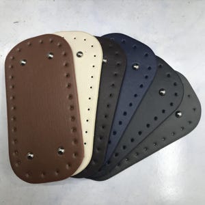 May include: A collection of oval-shaped bag bases in various colors, including brown, cream, black, navy, and gray. Each base has evenly spaced holes around the perimeter and metal rivets. These bases are designed for crafting.
