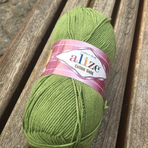 May include: A ball of green Alize Cotton Gold yarn rests on a wooden bench. The yarn is a vibrant, spring green color, and the label features the brand name and product details. This yarn is ideal for knitting and crochet projects.