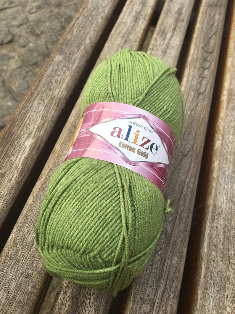 May include: A ball of green Alize Cotton Gold yarn rests on a wooden bench. The yarn is a vibrant, spring green color, and the label features the brand name and product details. This yarn is ideal for knitting and crochet projects.