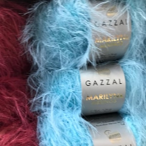 Puede incluir: Dos ovillos de hilo, uno es de color rojo burdeos oscuro y el otro es azul claro. Ambos ovillos son peludos y tienen la etiqueta de la marca Gazal Marilyn.
