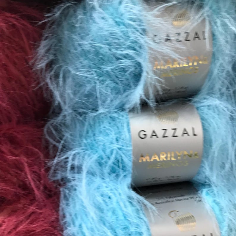 Puede incluir: Dos ovillos de hilo, uno es de color rojo burdeos oscuro y el otro es azul claro. Ambos ovillos son peludos y tienen la etiqueta de la marca Gazal Marilyn.