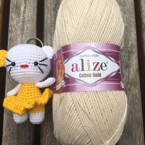 May include: A white and yellow crocheted cat keychain next to a large ball of cream-colored yarn. The yarn has a label that reads "alize Cotton Gold". The cat has black whiskers and a red mouth. The background is a wooden surface.