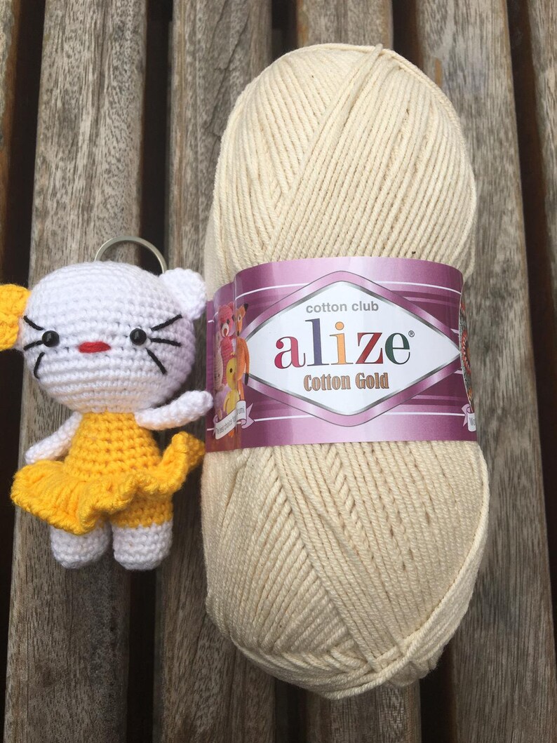 May include: A white and yellow crocheted cat keychain next to a large ball of cream-colored yarn. The yarn has a label that reads "alize Cotton Gold". The cat has black whiskers and a red mouth. The background is a wooden surface.