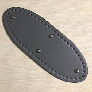 May include: An oval-shaped dark grey leather base for a bag, featuring a series of pre-punched stitching holes along the entire perimeter and four small, circular silver-toned metal feet attached to the surface for stability and protection.