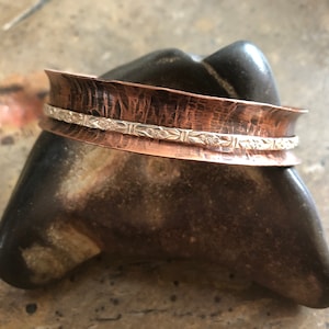 May include: A hammered copper cuff bracelet with a silver band featuring a floral design.