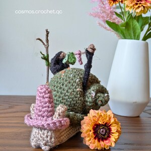 May include: A handmade crochet gnome with a pink hat and a green body, decorated with a miniature clothesline. The gnome is set on a wooden surface, with a flower and a vase of flowers in the background. The text "cosmos.crochet.qc" is visible.