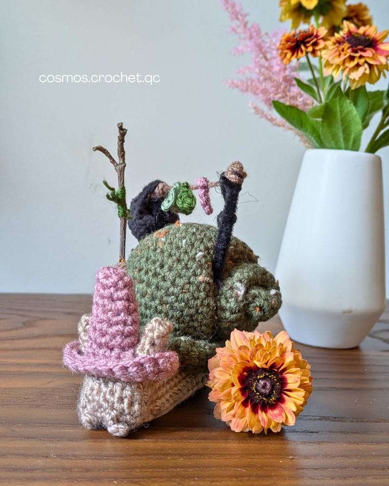 May include: A handmade crochet gnome with a pink hat and a green body, decorated with a miniature clothesline. The gnome is set on a wooden surface, with a flower and a vase of flowers in the background. The text "cosmos.crochet.qc" is visible.