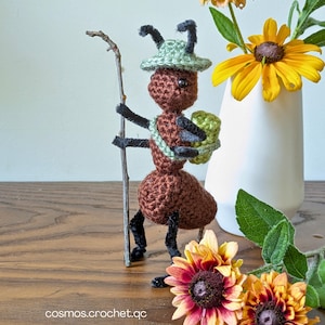 May include: A handmade crochet ant figurine, brown with black accents, wearing a green hat and carrying a green backpack and a stick. The ant is standing on a wooden surface, with a vase of yellow flowers in the background. The text "cosmos.crochet.qc" is visible.