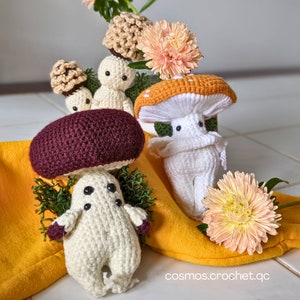 May include: Handmade crochet mushroom figures in shades of white, brown, and burgundy, arranged with peach-coloured flowers and green foliage on a yellow cloth. The image includes the text "cosmos.crochet.qc".