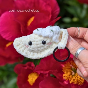 May include: A white crocheted dumpling charm with a black eye and a black ring attached. The charm is held by a hand against a background of red flowers.