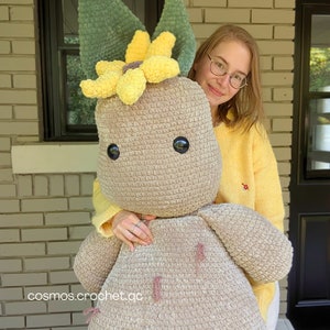May include: A crocheted plush toy shaped like a brown, furry creature with a yellow sunflower on top. The toy is being held by a person wearing a yellow sweater and glasses.