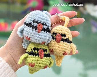 May include: Three crocheted bird keychains in different colors: light blue, yellow, and green. Each bird has black eyes and a red beak. The keychains are attached to a silver ring.