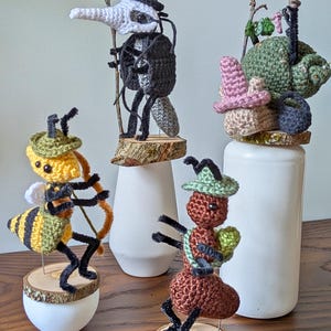 Könnte beinhalten: Eine Sammlung skurriler Häkelfiguren, darunter ein Pestdoktor, eine Schnecke, eine Ameise und eine Biene, jeweils mit einzigartigen Hüten und Accessoires. Die Figuren sind auf Holzsockeln montiert und auf weißen Sockeln ausgestellt. Der Text "cosmos.crochet.qc" ist sichtbar.