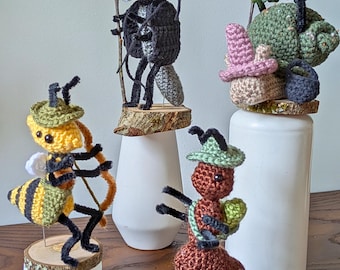 May include: A collection of whimsical crochet figures, including a plague doctor, a snail, an ant, and a bee, each with unique hats and accessories. The figures are mounted on wooden bases and displayed on white pedestals. The text "cosmos.crochet.qc" is visible.