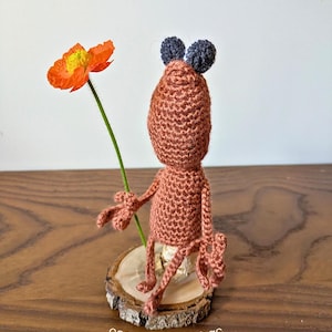 May include: A brown crocheted creature with two dark gray ears, standing on a circular wood slice base. The creature holds a single orange poppy flower with a yellow center. The text cosmos.crochet.qc is printed at the bottom of the image.