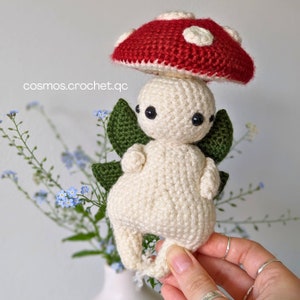 May include: A crocheted mushroom fairy with a red and white mushroom cap, green leaves, and a white body.