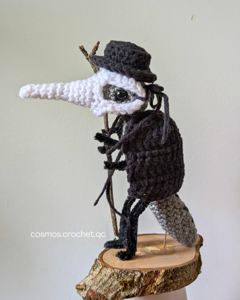 May include: A handcrafted crochet bird-like figure with a white beak and head, a black body, and a small black hat. It holds a twig and stands on a wooden base. The text "cosmos.crochet.qc" is visible.