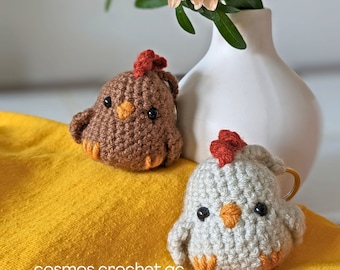 May include: Two crocheted chicken ornaments, one brown and one white, with red combs and beaks. The ornaments are sitting on a yellow surface.