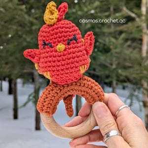 May include: A crochet bird-shaped baby rattle, predominantly red with yellow and orange detailing. The bird has embroidered features and is attached to a wooden ring. The text "cosmos.crochet.qc" is visible. The rattle is held in a hand.