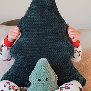 Op de afbeelding: Een kind houdt een groot groen gehaakt kerstboomkussen voor zijn gezicht. Een kleiner groen gehaakt kerstboomkussen met een glimlachend gezicht zit op zijn schoot. Het kind draagt een witte pyjama met een rood-groen kerstpatroon.
