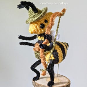 May include: A handmade crochet bee figurine, wearing a green hat and carrying a bow. The bee has black and yellow stripes and is mounted on a small wooden slice. The text "cosmos.crochet.qc" is visible in the background.