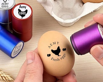 May include: A cylindrical purple handheld stamp is being used to imprint a black graphic of a chicken and the text AEMY Fresh eggs onto a brown egg. Several other colorful metal stamp cases in red, blue, and silver are visible nearby.