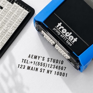 May include: A blue and black Trodat Printy 4911 typo self-inking stamp with a separate character set tray. The stamp has imprinted the text AEMY'S STUDIO, TEL:+1(555)1234567, and 123 MAIN ST NY 10001 in black ink on a white surface.