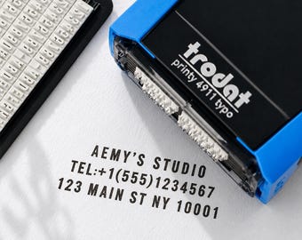 May include: A blue and black Trodat Printy 4911 typo self-inking stamp with a separate character set tray. The stamp has imprinted the text AEMY'S STUDIO, TEL:+1(555)1234567, and 123 MAIN ST NY 10001 in black ink on a white surface.