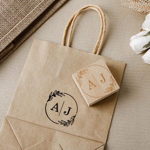 May include: A brown paper gift bag with twisted paper handles and a wooden stamp. The stamp features the initials "A J" inside a circular floral design. A similar design is printed on the bag.