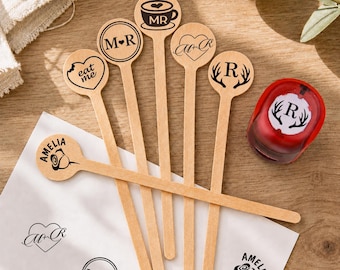 May include: Five light-colored wooden stirrers with circular tops featuring black laser-engraved designs including a coffee cup with MR, M+R, a heart with M+R, a heart with eat me, and antlers with R, alongside a red wax seal stamp with R and antlers.