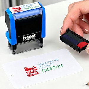 Op de afbeelding: Een blauwe en zwarte Trodat printy 4911 typo stempel met de tekst "CELEBRATING THE 250 YERS OF FREEDOM". De stempel staat naast een witte kaart met dezelfde tekst en een rood-wit-blauwe afbeelding.