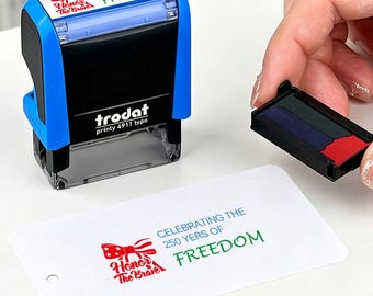 May include: A blue and black Trodat printy 4911 typo stamp with the text "CELEBRATING THE 250 YERS OF FREEDOM". The stamp is next to a white card with the same text and a red, white, and blue graphic.