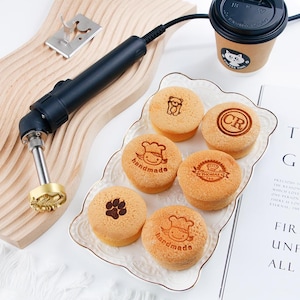 Puede incluir: Un hierro de marcar negro con un sello dorado junto a una tabla de cortar de madera. Varios pasteles pequeños y redondos están estampados con varios diseños y la palabra "handmade". También hay una taza de café con un logotipo de gato.