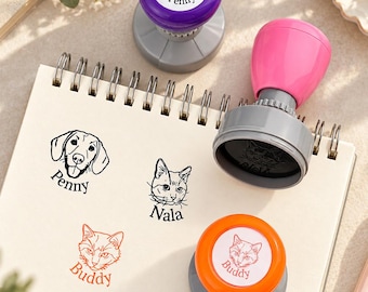 May include: Three circular self-inking stamps with purple, pink, and orange handles. The stamps feature custom pet portraits and names: Penny, Nala, and Buddy. The impressions are stamped on a spiral-bound notebook, showing black and orange ink designs.