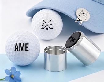 May include: Two white golf balls with dimpled surfaces, one featuring the text AME and the other with crossed golf clubs and the letters A, X, E. A light blue baseball cap features a circular metal ball marker with the text AMEY and crossed clubs. A silver metal container with a black lid is present.