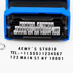 May include: A blue and black self-inking rubber stamp with a rectangular base. The stamp features raised white characters that read AEMY'S STUDIO, TEL: +1 (555) 1234567, and 123 MAIN ST NY 10001. A black ink impression of the same text is visible below.